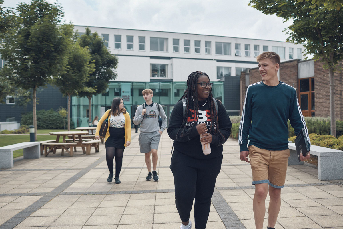 Four university students walk together through a campus courtyard, smiling and chatting. Two students are in the foreground, while two others walk behind them. The area is surrounded by trees, benches, and modern buildings with large windows.