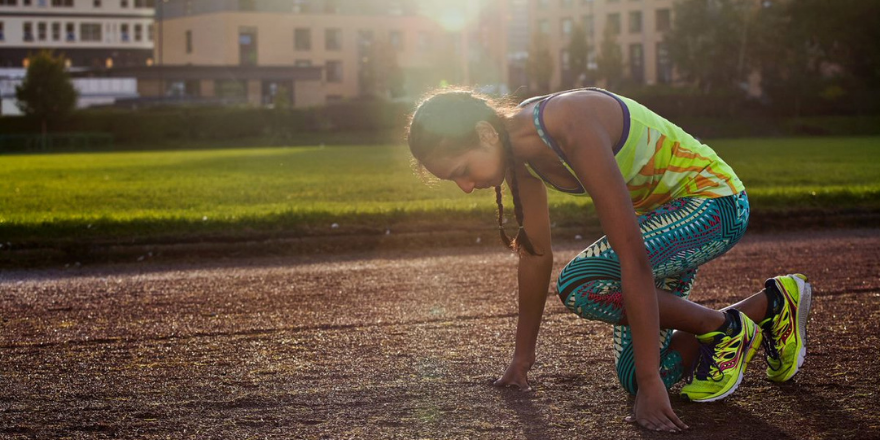 Girl with yellow sports top and blue leggings on crouches on running track in front of University