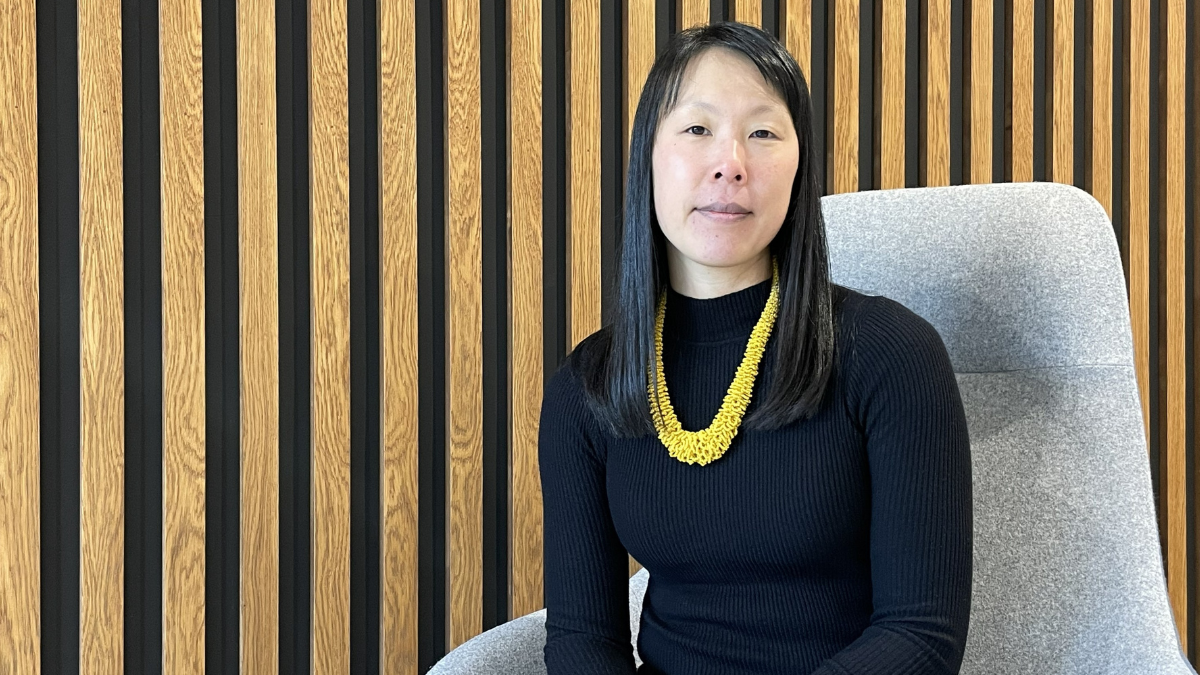 A female academic wearing a black top and a yellow necklace, sitting on a grey chair by a wall covered in wooden panels.