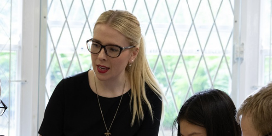 Blonde female in glasses with hair tied back and black top on speaks to students