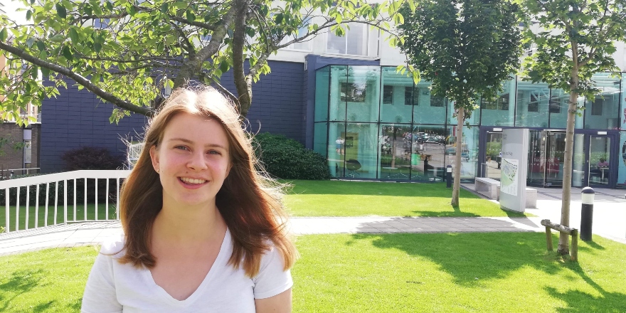 Girl with white top and shoulder length brown hair stands outside campus entrance
