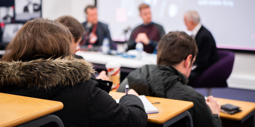 students in lecture theatre taking notes and listening to panel