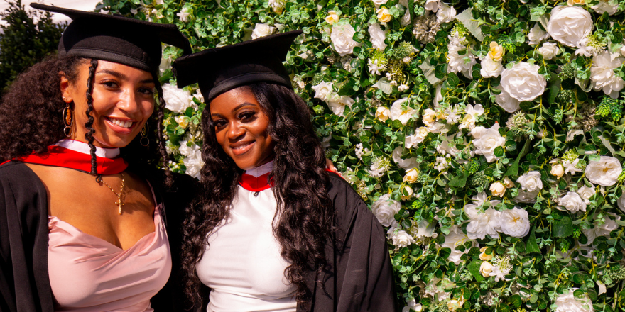 Two female Leeds Trinity graduates stand side by side with graduation caps on in front of a flower wall