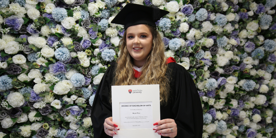 Blonde girl stands in front of flower wall with gown and graduation cap on holding degree certificate