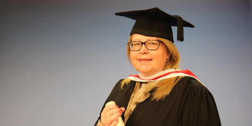 Female mature student with glasses poses for picture with graduation cap and gown, holding scroll