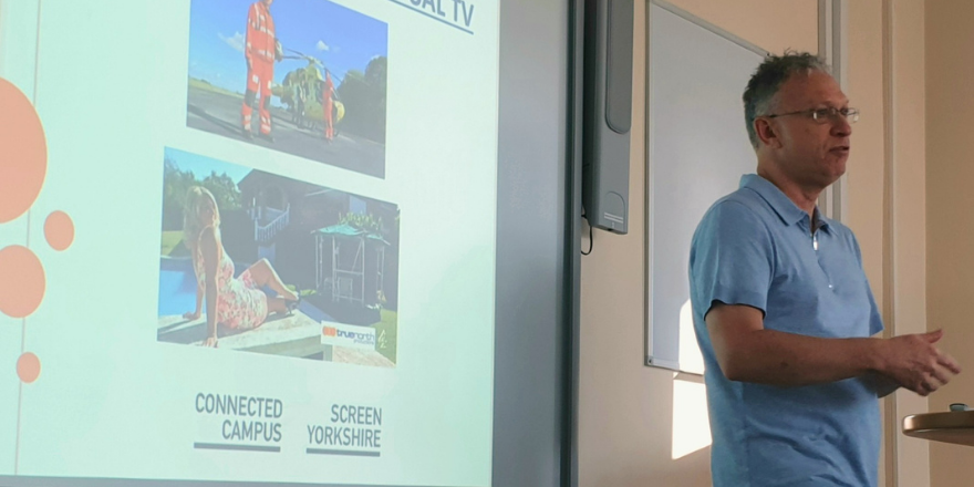 Man with grey hair and sky blue t-shirt stands presenting in front of screen