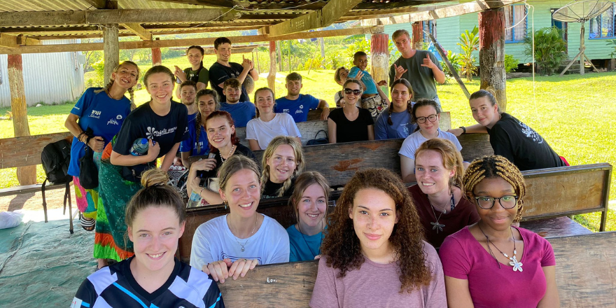 Large group of female and male students sit outside under wooden hut