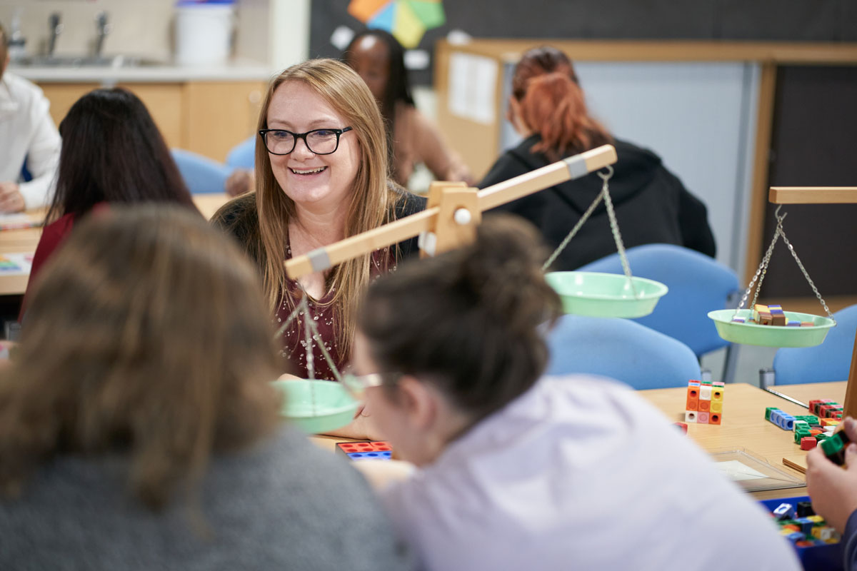 A student smiles while working with classroom resources, including scales and colourful counting blocks, during a group activity.